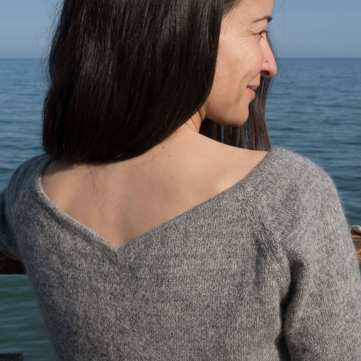 Alpaka Pullover Mia mit femininem V-Ausschnitt hinten, getragen von dunkelhaarigem Model das aufs Meer blickt